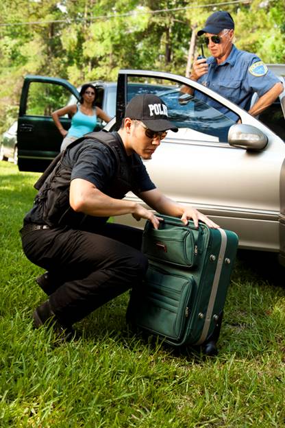 A man in a "POLICE" cap kneels, placing a green suitcase in a car trunk. Two other people stand nearby on the grass.